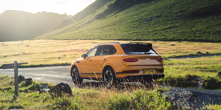 Bentley Kuala Lumpur Bentley Bentayga S SUV rear three quarter in Sunburst Gold paint on a road surrounded by green hills