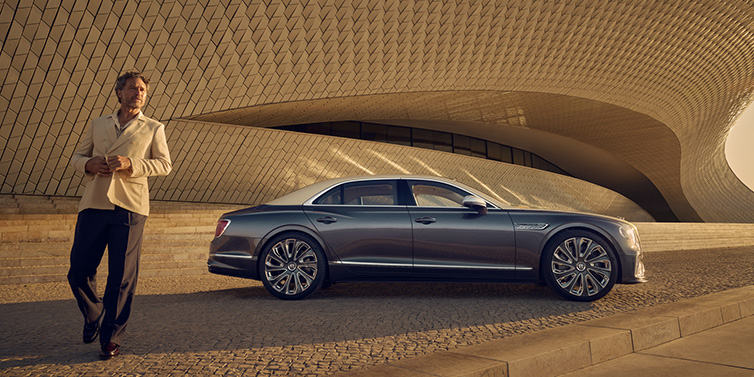 Bentley Kuala Lumpur Bentley Flying Spur Mulliner sedan in Rose Gold over Magnetic duotone paint parked by a curved wall with person in suit jacket standing on the left